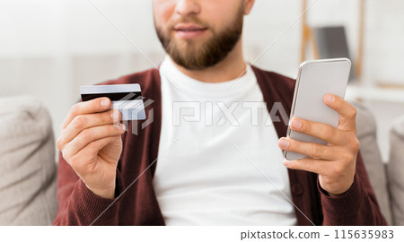 A man holding a smart phone and a credit card in his hands, showcasing the action of making a digital payment transaction. The mans face is focused and intent on the screen of the phone 115635983