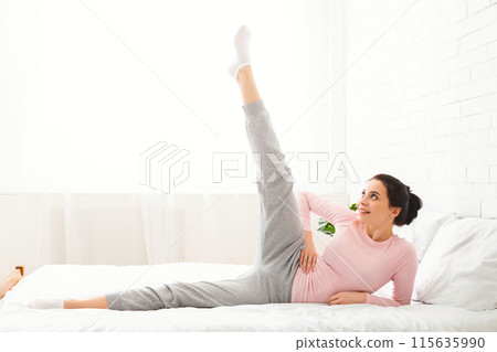 A woman is practicing a morning stretch routine on her bed in a well-lit bedroom, dressed in comfortable loungewear. She appears relaxed and focused. A woman is practicing a morning stretch routine on her bed in a well-lit bedroom, dressed in comfortable loungewear. She appears relaxed and focused. 115635990