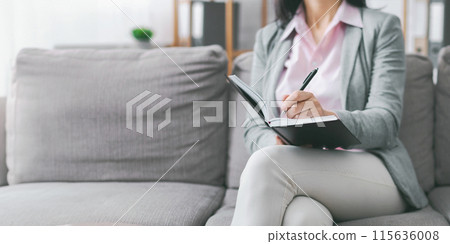 A woman sits on a gray couch in a living room. She is writing in a black notebook with a pen in her right hand. The scene is calm and serene, with the woman focused on her writing, copy space A woman sits on a gray couch in a living room. She is writing in a black notebook with a pen in her right hand. The scene is calm and serene, with the woman focused on her writing, copy space 115636008