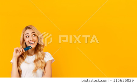 A young woman with long, blonde hair is holding a credit card to her mouth and thinking. She is wearing a white t-shirt and has a bright, cheerful expression on her face, copy space 115636029