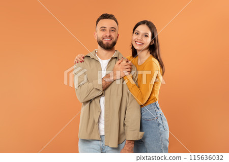A cheerful couple is smiling at the camera while standing against an orange backdrop. They are casually dressed in a beige jacket and a mustard yellow top with jeans. 115636032