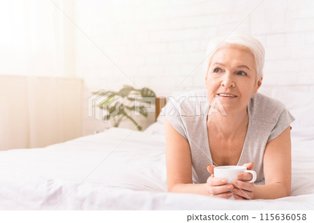 Old woman is laying on top of a neatly made bed, holding a cup in her hands. She is relaxed and comfortable, enjoying a moment of rest, copy space Old woman is laying on top of a neatly made bed, holding a cup in her hands. She is relaxed and comfortable, enjoying a moment of rest, copy space 115636058