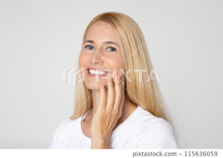 This image features a blonde woman with a white t-shirt smiling while she touches her face with her hand. The background is a plain white wall. Her blue eyes are bright and friendly. This image features a blonde woman with a white t-shirt smiling while she touches her face with her hand. The background is a plain white wall. Her blue eyes are bright and friendly. 115636059