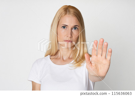 A blonde woman wearing a white t-shirt is looking directly at the camera with a serious expression. She is holding her right hand up, palm facing forward, in a clear stop gesture 115636063