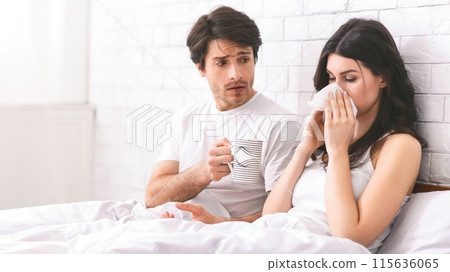 A man with a worried expression holds a mug of tea while a sick woman blows her nose with a tissue in bed. The couple are in a bedroom with a white brick wall. 115636065