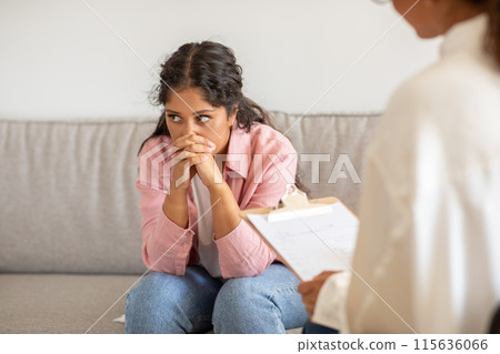 A young woman sits on a couch in a counseling session, looking down and holding her hands together in her lap. She is clearly troubled, while the therapist sits across from her 115636066