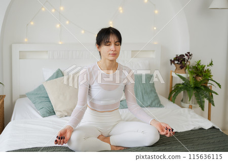 Yoga mindfulness meditation. Young healthy woman practicing yoga in bedroom at home. Woman sitting in lotus pose on bed meditating smiling relaxing indoor. Girl doing breathing practice. Yoga at home Yoga mindfulness meditation. Young healthy woman practicing yoga in bedroom at home. Woman sitting in lotus pose on bed meditating smiling relaxing indoor. Girl doing breathing practice. Yoga at home 115636115