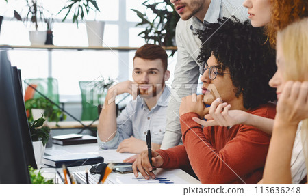 A group of four multiethnic coworkers is gathered around a computer monitor, focused on a collaborative work discussion. The office setting is casual and creative 115636248