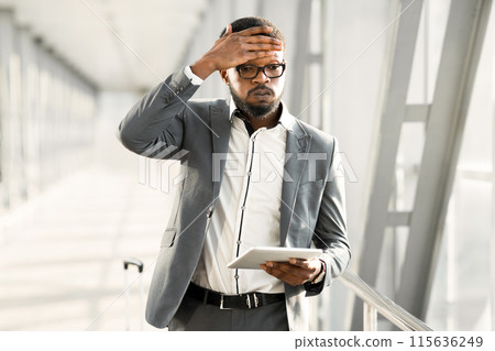 Distressed black businessman wearing eyeglasses holding a tablet while standing in a bright, modern hallway, touching his head 115636249