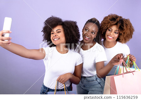 Three Cheerful Young African American Women On Shopping Taking Selfies Using Smartphone, Posing Together Holding Colorful Shopper Bags, Advertising And Enjoying Sales Season Over Purple Background 115636259
