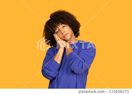 A young man with curly hair is wearing a blue sweater and posing as if he is sleeping, with hands together under his cheek. He stands in front of a vibrant orange background 115636273