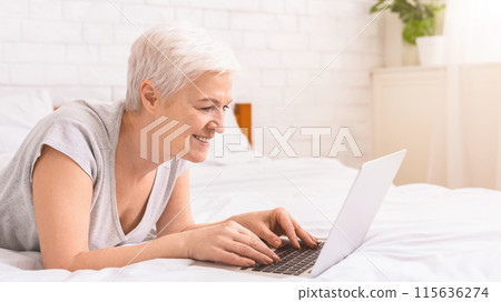 Senior woman is laying on a bed, fully engrossed in using a laptop computer. She is positioned comfortably with the laptop in front of her, typing and browsing, copy space Senior woman is laying on a bed, fully engrossed in using a laptop computer. She is positioned comfortably with the laptop in front of her, typing and browsing, copy space 115636274