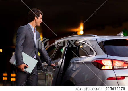 Handsome young businessman wearing expensive grey suit manager with laptop computer, phone and documents in his hand getting into luxury car in the evening, going home after busy day 115636276