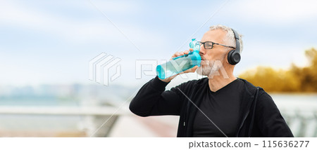 Elderly handsome sportsman wearing black sportswear using wireless headphones drinking water, standing on walking bridge, looking at copy space. Senior man have outdoor workout, web-banner 115636277