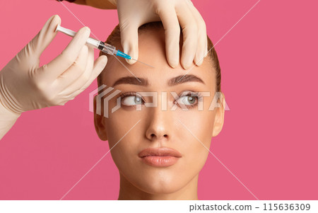 A woman receives a forehead injection treatment from a medical professional wearing gloves. The image is taken against a pink background. The womans expression is relaxed and she is looking upwards. A woman receives a forehead injection treatment from a medical professional wearing gloves. The image is taken against a pink background. The womans expression is relaxed and she is looking upwards. 115636309
