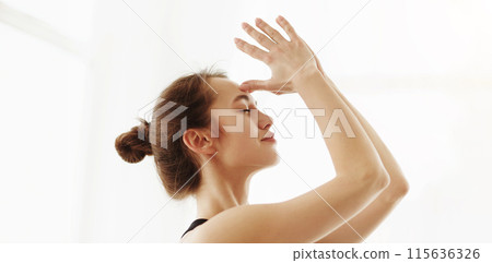 A serene young woman with her hair tied up in a bun is performing a yoga pose with a delicate hand gesture, her face exuding calmness. Sunlight gently illuminates her, closeup 115636326