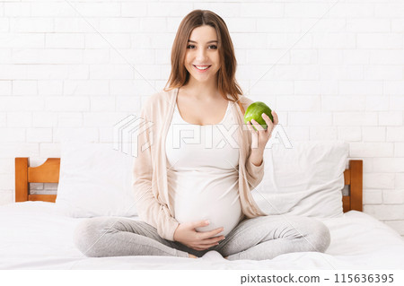 A pregnant woman sitting on a bed, holding a green apple in her hands. She appears calm and content as she gazes at the apple in her hands. A pregnant woman sitting on a bed, holding a green apple in her hands. She appears calm and content as she gazes at the apple in her hands. 115636395