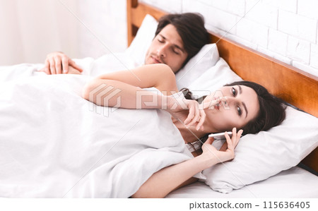 A woman is lying in bed next to a sleeping man, holding a phone to her ear while whispering into it with her finger on her lips. The man appears to be asleep, unaware of secretive phone call. 115636405