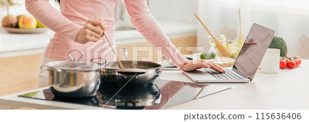 A woman in a pink shirt stirs food in a frying pan while referencing her laptop in a bright, modern kitchen filled with fresh ingredients, cropped 115636406