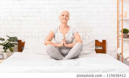 A senior woman with a gentle smile is sitting cross-legged on a bed in a sunlit bedroom, her hands resting on her knees in a meditative pose A senior woman with a gentle smile is sitting cross-legged on a bed in a sunlit bedroom, her hands resting on her knees in a meditative pose 115636412