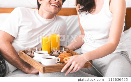 A woman is serving her partner breakfast in bed. She is holding a wooden tray with coffee, orange juice, croissants, and granola. The man is smiling and looking at the woman, cropped 115636435