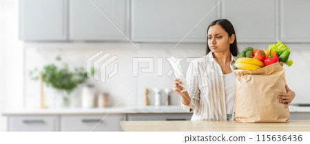 Indian woman stands in her kitchen, looking at a receipt in her hand with a shocked expression. She holding a paper bag filled with fresh goods, copy space 115636436