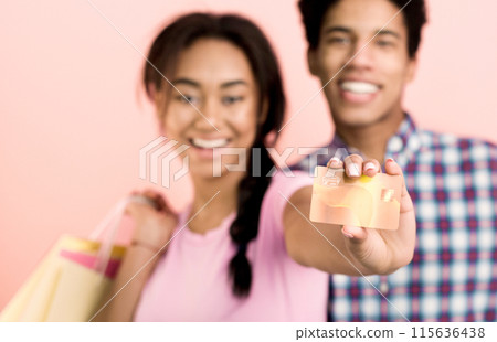 A young African American couple stands together, holding a credit card in front of a vibrant pink background. They both appear focused and engaged, possibly discussing financial decisions 115636438