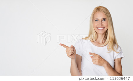 A woman with long blonde hair is smiling and pointing to the copy space in front of a plain white background. She is wearing a simple white t-shirt and has a friendly expression on her face. 115636447