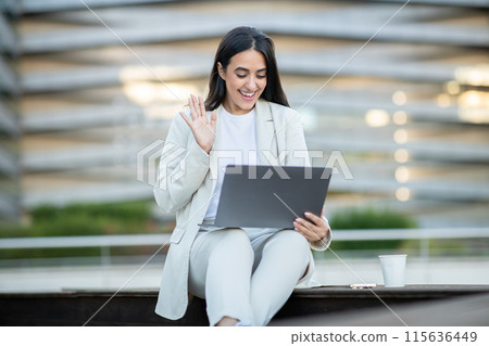 A young woman in a white suit sits on a bench outside a modern office building. She is using a laptop and waving at the camera, suggesting she is engaged in a video call A young woman in a white suit sits on a bench outside a modern office building. She is using a laptop and waving at the camera, suggesting she is engaged in a video call 115636449