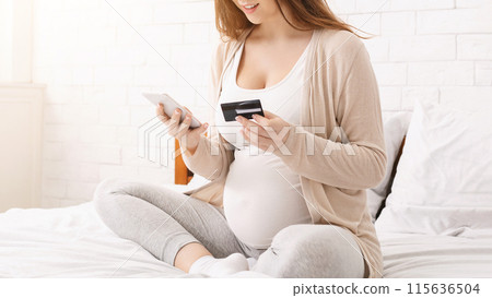 A happy pregnant woman sits in bed on a bright morning, using her smartphone to shop online while holding a credit card. She is dressed comfortably in light casual clothing, cropped A happy pregnant woman sits in bed on a bright morning, using her smartphone to shop online while holding a credit card. She is dressed comfortably in light casual clothing, cropped 115636504