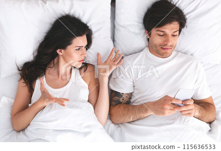 An image depicting a couple lying in bed. The woman is looking at the man, with her hand outstretched, seemingly trying to get his attention. He is ignoring her and looking at his phone 115636533