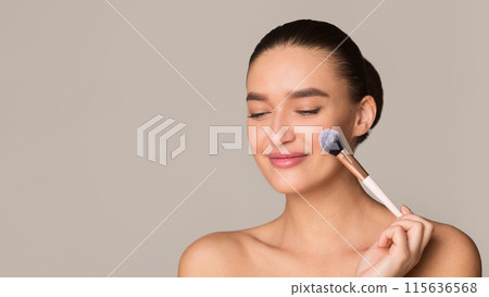 A woman is applying makeup to her face using a soft makeup brush. She carefully sweeps the brush across her cheeks and forehead, blending in foundation and blush to achieve a flawless look. 115636568