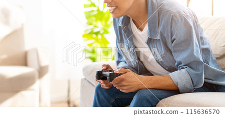 A teen person, wearing a blue denim shirt and jeans, sits on a couch in a living room. They are intently playing a video game, holding a black controller in their hands, cropped A teen person, wearing a blue denim shirt and jeans, sits on a couch in a living room. They are intently playing a video game, holding a black controller in their hands, cropped 115636570