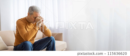 A man sits on a couch in his living room, his hands covering his face as he looks down. He appears to be in distress or feeling overwhelmed with emotion, possibly sadness or depression, copy space 115636620