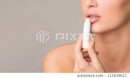 A woman applying lip balm to her lips in an indoor setting. The scene is a close-up shot focusing on the lower half of her face, emphasizing the action of moisturizing her lips, cropped, copy space 115636621