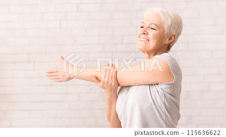 A joyful elderly woman with short white hair is engaging in a light stretching routine, extending her arms across her chest and smiling widely 115636622