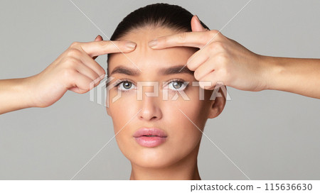 A woman with tied-back hair is using her fingers to squeeze a pimple on her forehead. The setting appears bright and indoor, suggesting a well-lit room. She has a calm yet focused expression A woman with tied-back hair is using her fingers to squeeze a pimple on her forehead. The setting appears bright and indoor, suggesting a well-lit room. She has a calm yet focused expression 115636630