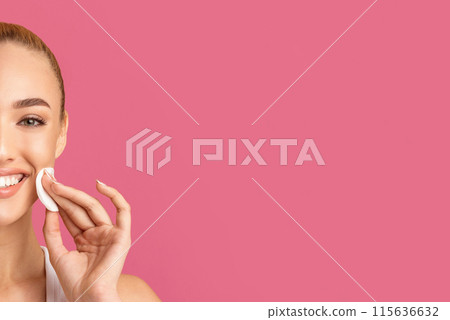 A young woman with long blonde hair is smiling and applying a cotton pad to her cheek. She is standing in front of a solid pink background. Her expression is happy and relaxed, copy space 115636632