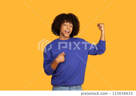 A young man with a dark curly and a big smile stands against a yellow backdrop, flexing his bicep while pointing at himself with his other hand. He appears confident and proud of his strength 115636633