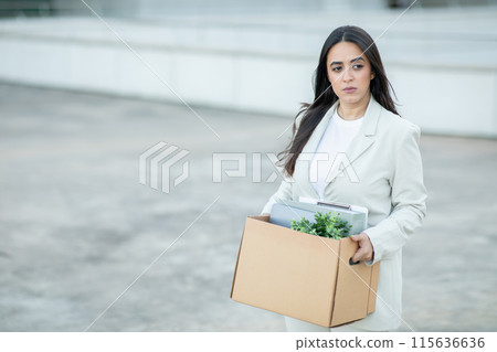 A young woman, dressed in a white blazer and a white shirt, walks away from a building, carrying a cardboard box filled with personal items. She has a serious expression on her face. 115636636