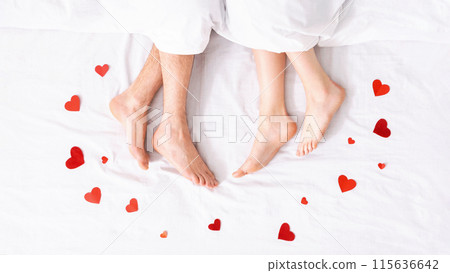 An overhead shot of two bare feet resting in a white bed with red paper hearts scattered around them. The feet are presumably from a couple in bed together, suggesting a romantic setting. 115636642