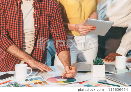 Diverse group of individuals standing, engaged in discussion, looking at tablet on table in modern office setting. 115636654