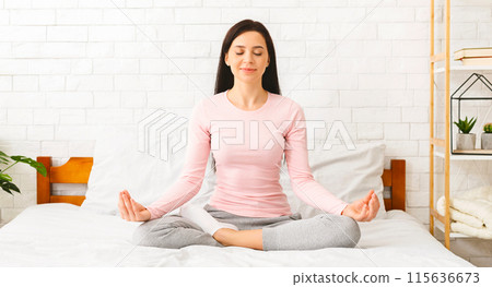 A woman is seen sitting in a lotus position on a neatly made bed, with her legs crossed and hands resting on her knees. She appears calm and focused, embodying a sense of mindfulness and relaxation. 115636673