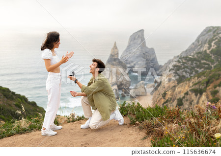 Happy young man making proposal to his surprised girlfriend, standing on knee with beautiful coastline view, copy space. Special romance marriage outdoors Happy young man making proposal to his surprised girlfriend, standing on knee with beautiful coastline view, copy space. Special romance marriage outdoors 115636674