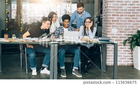 A group of diverse students are seated around a large conference table in a modern library. They appear engaged in a meeting or discussion, with papers and laptops scattered around the table. A group of diverse students are seated around a large conference table in a modern library. They appear engaged in a meeting or discussion, with papers and laptops scattered around the table. 115636675