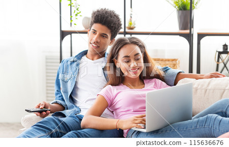 Black couple relaxes on a couch in their home. The girl is using a laptop computer while the guy watches television with a remote control in his hand. They both appear happy and content. 115636676