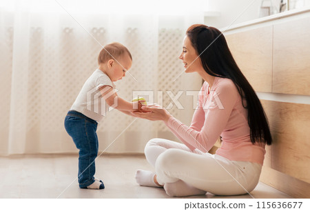 A mother sits on the floor, smiling as she interacts with her baby, who is standing and handing her a toy in a well-lit living room. 115636677