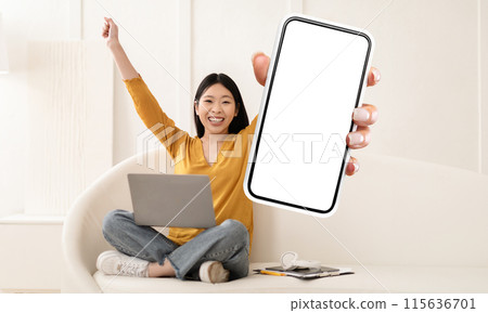 A cheerful young asian woman, dressed in a casual yellow top and jeans, sits cross-legged on a cozy white sofa with her laptop, enthusiastically presenting a smartphone with a blank screen 115636701