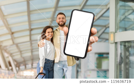 A cheerful man and woman stand together in an airport terminal, with the man extending his arm towards the camera, showing a smartphone with a blank screen A cheerful man and woman stand together in an airport terminal, with the man extending his arm towards the camera, showing a smartphone with a blank screen 115636719