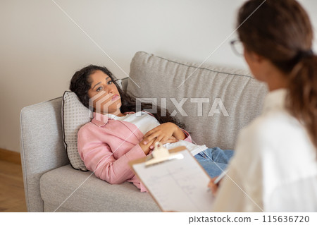 A young woman lies on a couch during a therapy session. She is dressed in a pink jacket and jeans. She appears to be sad or anxious. A therapist sits in front of her and appears to be listening 115636720